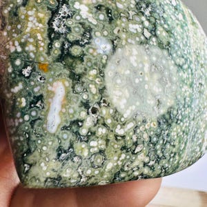 May include: Close-up of a polished gemstone with a mottled pattern. The stone features a mix of green, white, and yellow hues, with a speckled texture. The surface has a smooth, rounded shape, showcasing natural variations.