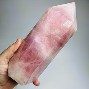 May include: A large, polished rose quartz crystal tower with a hexagonal shape and a pointed top. The stone features a translucent, pale pink color with natural internal fractures, cloudy white inclusions, and smooth, faceted surfaces.