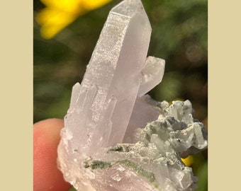 May include: A cluster of clear quartz crystals with a purple hue, some of the crystals are pointed and others are more rounded. The crystals are set in a matrix of green and white material.