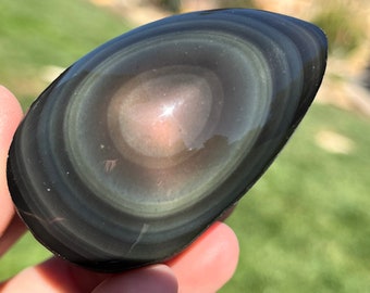 May include: A black obsidian sphere with a swirling, iridescent pattern. The sphere is smooth and polished, with a dark, almost black, background. The iridescent pattern is made up of shades of green, blue, and purple.