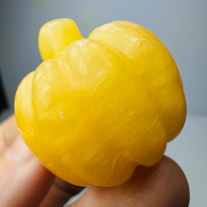 May include: A small, bright yellow carved stone figurine shaped like a pumpkin. The object features a smooth, polished surface with vertical ridges and a small, rounded stem at the top, showcasing a translucent, vibrant golden-yellow color throughout.