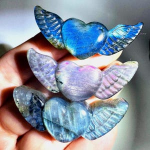 May include: Three carved labradorite stone ornaments shaped like hearts with wings. The top heart is vibrant blue, the middle heart is a soft lavender purple, and the bottom heart is a muted blue-grey. Each piece features detailed wing carvings.