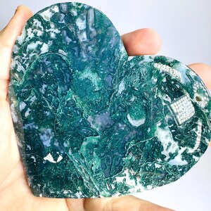 May include: A heart-shaped, polished stone with a mix of deep green and white patterns. The stone has a natural, textured surface and is held in a hand. The stone's color and shape suggest it could be used as a decorative item or a paperweight.