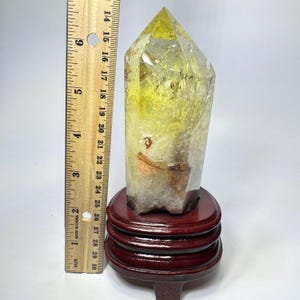 May include: A yellow and clear quartz crystal point with internal fractures and inclusions, mounted on a dark brown circular wooden stand. A wooden ruler is placed vertically next to the crystal, showing a height of approximately 5.5 inches.