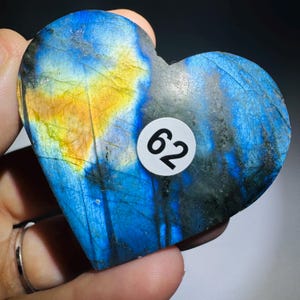 May include: A heart-shaped labradorite stone featuring a polished surface with iridescent blue and yellow flashes. A small circular white sticker with the black number 62 is affixed to the center of the stone, which displays natural mineral inclusions.