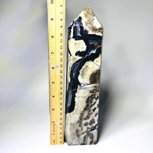 May include: A polished stone obelisk, approximately 9 inches tall, featuring a striking pattern of beige, black, and white veining. The obelisk has a rectangular base and a pointed top. A wooden ruler is placed next to the obelisk.