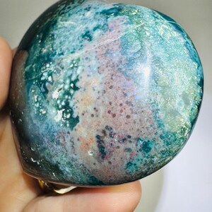 May include: A polished, rounded stone with a mottled pattern of teal, green, and reddish-brown hues. The stone's surface has a glossy sheen, and the colors blend together in a natural, organic design. The stone is held in a hand.