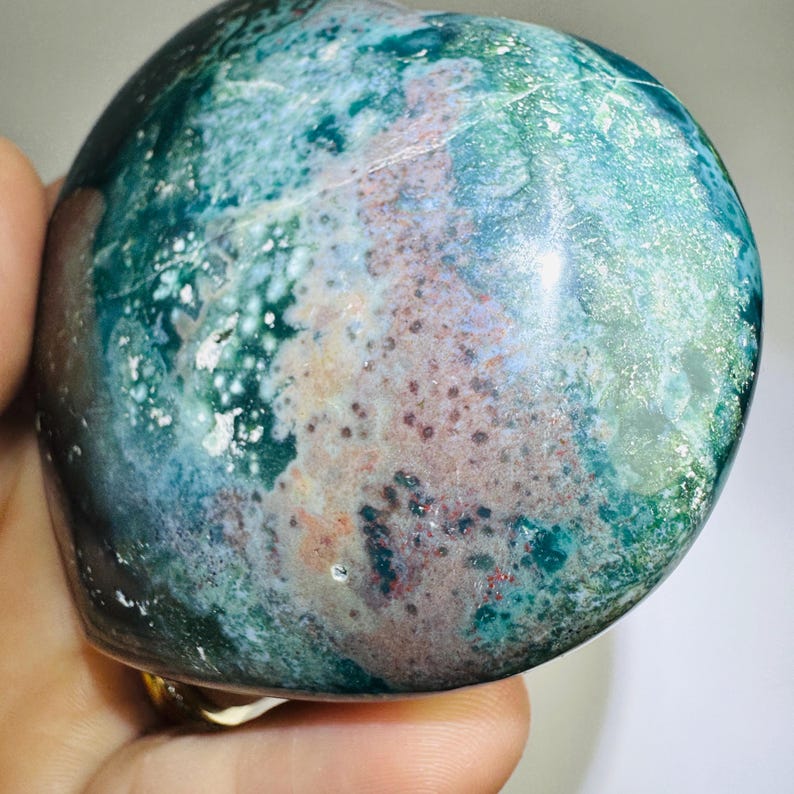 May include: A polished, rounded stone with a mottled pattern of teal, green, and reddish-brown hues. The stone's surface has a glossy sheen, and the colors blend together in a natural, organic design. The stone is held in a hand.