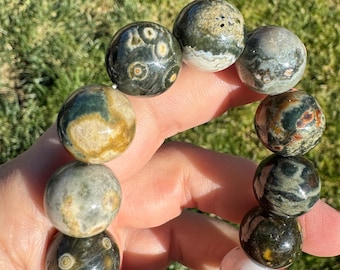 May include: A beaded bracelet featuring large, round gemstones in shades of green, brown, and cream. The stones have unique patterns and textures, creating a natural and earthy aesthetic. The bracelet is held in a hand, showcasing the polished finish of the beads.
