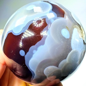 May include: A polished spherical stone featuring a smooth surface with deep reddish-brown and translucent light blue circular patterns. The orb displays distinct banded layers and cloudy inclusions that create a marbled effect across the entire sphere.