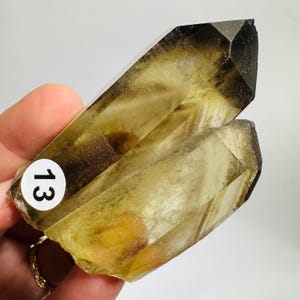 May include: A translucent, golden-brown quartz crystal cluster. The crystal has a dark brown tip and a sticker with the number 13 on it. The crystal is held in a hand.