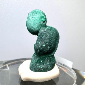 Op de afbeelding: Een levendig groen malachiet mineraal specimen, gevormd in een unieke vorm, rust op een witte basis. De malachiet toont een textuur met verschillende groentinten. Het specimen wordt gepresenteerd tegen een neutrale achtergrond.