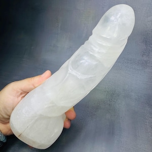 Peut inclure: Une sculpture en forme de phallus taillée dans du cristal de quartz clair et translucide. L'objet présente une surface lisse et polie avec des inclusions internes naturelles et une extrémité arrondie, révélant la structure cristalline.