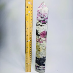 May include: A polished crystal tower featuring a mix of white, grey, black, and vibrant pink mineral inclusions. A yellow wooden ruler is placed vertically next to the crystal, indicating a total height of approximately 11.5 inches.