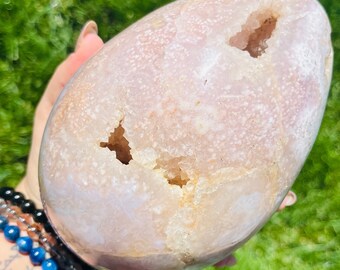 Puede incluir: Un huevo de geoda de ágata rosa claro grande con un exterior liso y pulido y varias aberturas que revelan un interior cristalino blanco.