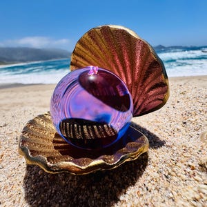 May include: A spherical glass orb with a translucent purple and blue iridescent finish resting inside a decorative gold-toned metal clam shell. The shell features a textured, ridged surface with a metallic sheen, cradling the smooth, reflective glass sphere.