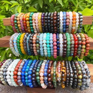 May include: A collection of colorful gemstone bracelets stacked on a wooden display. The bracelets are made of various types of stones, including amethyst, rose quartz, tiger's eye, and turquoise.