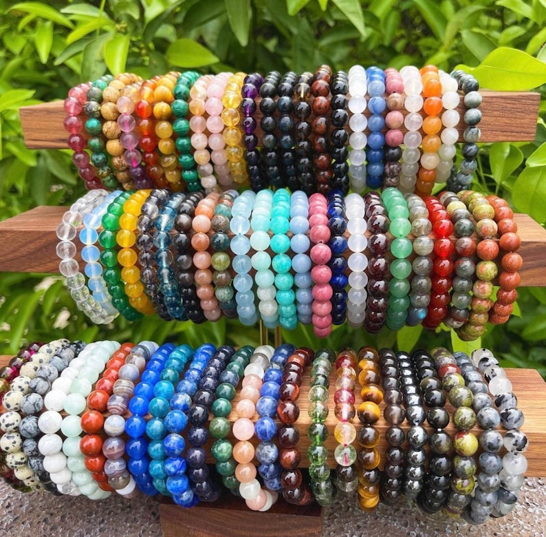 May include: A collection of colorful gemstone bracelets stacked on a wooden display. The bracelets are made of various types of stones, including amethyst, rose quartz, tiger's eye, and turquoise.