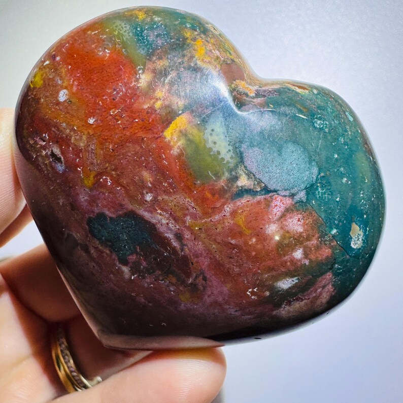 May include: A polished heart-shaped stone featuring a mottled pattern of deep forest green, earthy red, ochre yellow, and dark brown tones with a smooth, glossy surface that reflects light across its varied mineral inclusions.