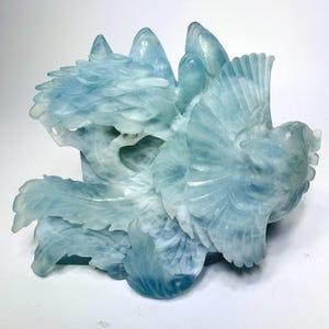 May include: A translucent light blue carved stone sculpture depicting a bird with spread wings and detailed plumage, positioned next to a stylized tree structure with rounded foliage and mountain-like peaks in the background.
