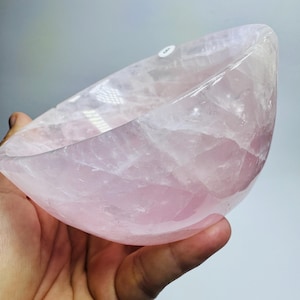 May include: A polished bowl carved from translucent pale pink rose quartz. The bowl features a smooth, curved interior and exterior surface with visible natural crystalline fractures and internal inclusions throughout the light pink stone material.
