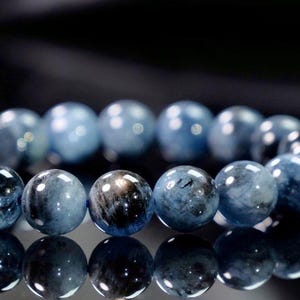 May include: A bracelet composed of polished, spherical blue beads with dark, marbled inclusions. The beads exhibit a glossy, reflective surface with varying shades of deep and light blue, arranged in a continuous circular strand on a dark surface.