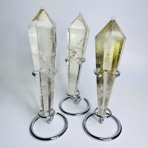 May include: Three clear quartz crystal points with a yellow hue, each mounted on a silver metal stand. The crystals are faceted and taper to a point. The stands have a circular base and a ring to hold the crystal.