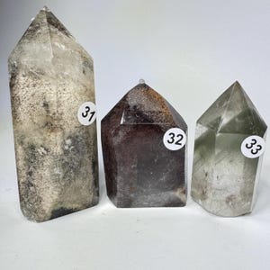 May include: Three polished crystal points standing upright. The tallest, labeled 31, is clear with grey and white inclusions. The middle, labeled 32, is dark brown with a reddish-gold top. The third, labeled 33, is translucent with green hues.