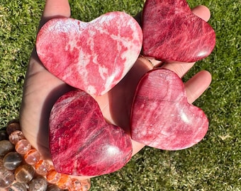 May include: Four red and white heart-shaped polished stones. The stones have a marbled appearance and are smooth to the touch.