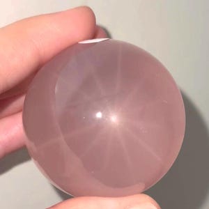 Peut inclure: Une sphère en quartz rose polie et lisse, présentant une teinte rose pâle translucide. La surface de la pierre révèle un motif d'astérisme subtil en forme d'étoile sous la lumière, reflétant un éclat doux sur sa forme arrondie et symétrique.