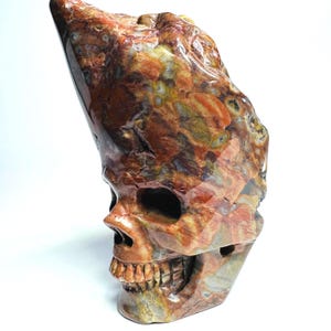 May include: A polished stone carving in the shape of a human skull. The material features a mottled pattern of earthy tones including deep red, burnt orange, ochre, and cream, with a smooth, glossy finish across the entire surface structure.