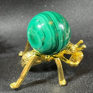 May include: A green malachite sphere on a gold metal stand with three legs. The sphere has a swirling pattern of green and gold.