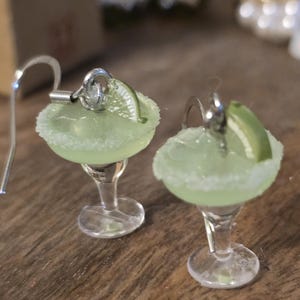 May include: A pair of margarita earrings. Each earring is a miniature margarita glass with a light green liquid, a salt-rimmed edge, and a lime wedge. The earrings have silver-colored fish hook ear wires.