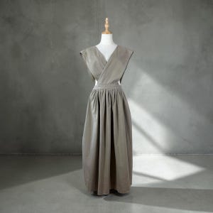 May include: A taupe-colored dress displayed on a mannequin. The dress features a V-neckline, wrap-around bodice, and a full, pleated skirt. The dress is floor-length and appears to be made of a soft, flowing fabric. The background is a neutral gray.