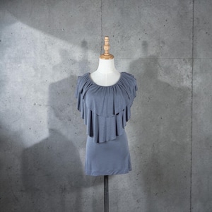 May include: A dusty blue top with a layered ruffle design on a mannequin. The top has a round neckline and a fitted silhouette. The mannequin is on a metal stand against a gray concrete wall.