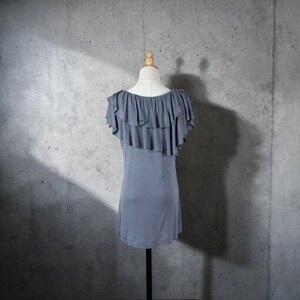 May include: A gray, short-sleeved top with a ruffled neckline and shoulder detail. The top is displayed on a mannequin against a gray concrete background. The ruffles cascade down the left shoulder, adding a touch of elegance to the simple design.
