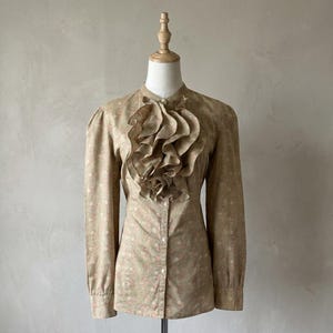 Puede incluir: Una blusa beige con estampado floral y un gran cuello con volantes. La camisa de manga larga tiene una botonadura frontal y se muestra en un maniquí. La tela parece ligera, adecuada para climas cálidos.