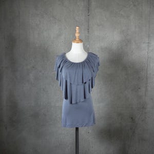 May include: A dusty blue top with layered ruffles around the neckline and shoulders. The top is displayed on a mannequin against a gray background. The garment has a simple, fitted silhouette.