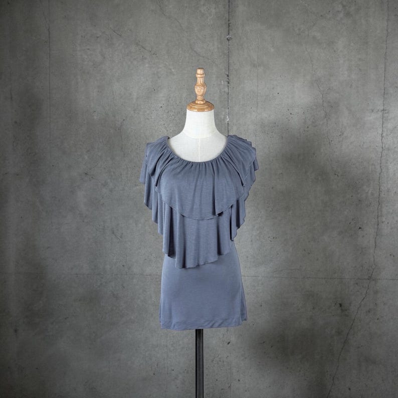 May include: A dusty blue top with layered ruffles around the neckline and shoulders. The top is displayed on a mannequin against a gray background. The garment has a simple, fitted silhouette.