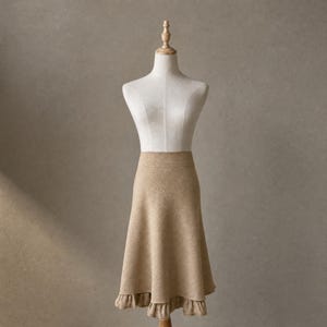May include: A beige skirt with a flared silhouette and a ruffled hem is displayed on a mannequin. The skirt has a high waist and a soft, textured appearance. The mannequin is set against a neutral background.