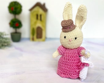 May include: A handmade crochet bunny doll wearing a pink dress and a brown hat. The bunny is holding a smaller white bunny. The doll is cream-colored with pink accents.