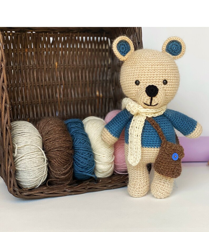 Pode incluir: Um urso de pel&uacute;cia de croch&ecirc; usando um su&eacute;ter azul, cachecol creme e bolsa marrom com um bot&atilde;o azul. Ao lado, uma cesta de vime cheia de fios em v&aacute;rias cores: creme, marrom, azul e rosa.