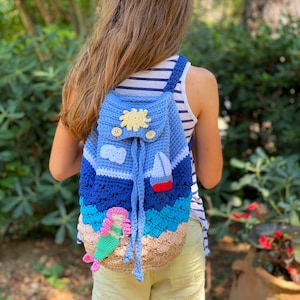 May include: A blue crochet backpack with a beach theme. The backpack features a sun, clouds, a sailboat, and a mermaid. The bag has a drawstring closure and wooden buttons. The backpack is worn by a person.