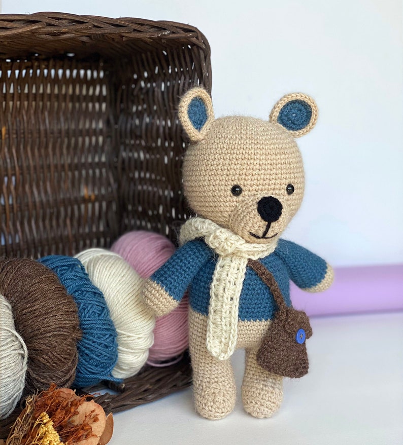 Pode incluir: Um urso de pel&uacute;cia de croch&ecirc; feito &agrave; m&atilde;o com corpo bege, detalhes azuis e um cachecol creme. O urso usa um su&eacute;ter azul e uma bolsa marrom com um bot&atilde;o azul. Meadas de fios de v&aacute;rias cores e uma cesta de vime ao fundo.