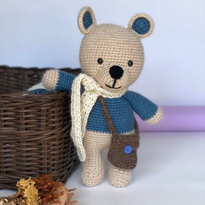 Pode incluir: Um urso de pel&uacute;cia de croch&ecirc; feito &agrave; m&atilde;o, com cabe&ccedil;a e corpo bege, bra&ccedil;os e detalhes azuis e um cachecol creme. O urso tem uma pequena bolsa marrom com um bot&atilde;o azul e um nariz e boca costurados em preto.