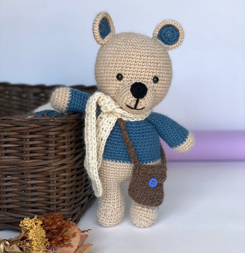 Pode incluir: Um urso de pel&uacute;cia de croch&ecirc; feito &agrave; m&atilde;o, com cabe&ccedil;a e corpo bege, bra&ccedil;os e detalhes azuis e um cachecol creme. O urso tem uma pequena bolsa marrom com um bot&atilde;o azul e um nariz e boca costurados em preto.