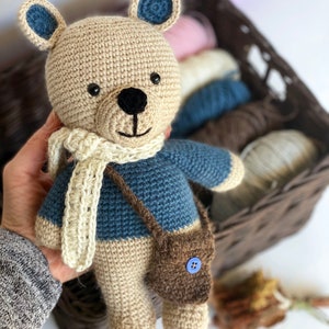 Pode incluir: Um urso de pel&uacute;cia de croch&ecirc; feito &agrave; m&atilde;o com corpo bege, su&eacute;ter azul e uma pequena bolsa marrom. O urso tem um cachecol creme e detalhes azuis nas orelhas. Ele segura uma pequena bolsa marrom com um bot&atilde;o azul.