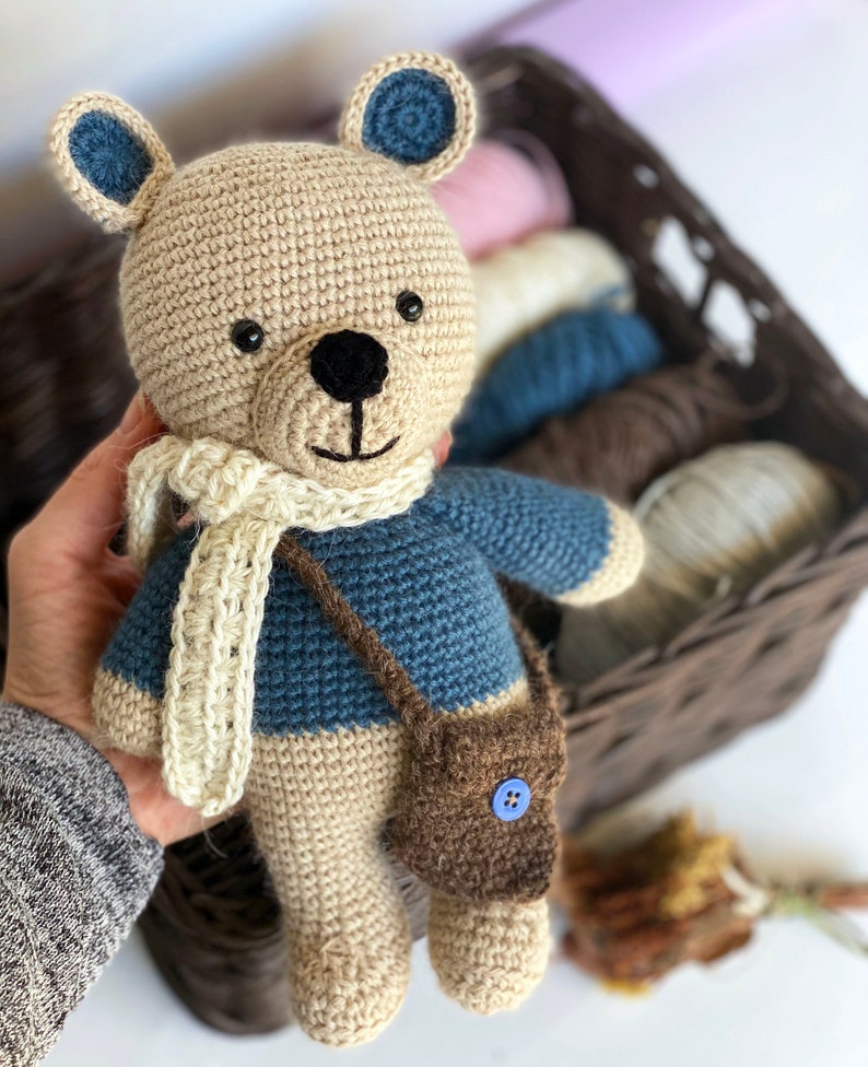 Pode incluir: Um urso de pel&uacute;cia de croch&ecirc; feito &agrave; m&atilde;o com corpo bege, su&eacute;ter azul e uma pequena bolsa marrom. O urso tem um cachecol creme e detalhes azuis nas orelhas. Ele segura uma pequena bolsa marrom com um bot&atilde;o azul.