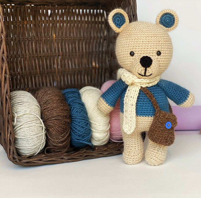 Pode incluir: Um urso de pel&uacute;cia de croch&ecirc; usando um su&eacute;ter azul, cachecol creme e uma bolsa marrom com um bot&atilde;o azul. O urso tem nariz e olhos pretos e detalhes azuis nas orelhas. Uma cesta de vime cont&eacute;m novelos de l&atilde; em v&aacute;rias cores.