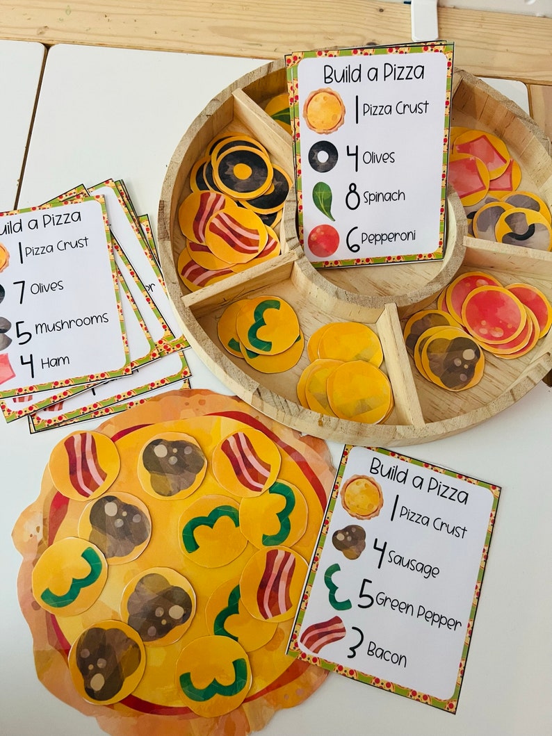 K&ouml;nnte beinhalten: Ein Satz bedruckbarer Karten zum Thema Pizza f&uuml;r ein Lernspiel f&uuml;r Kinder. Die Karten zeigen verschiedene Pizzabel&auml;ge mit Zahlen, die die Menge jedes Belags angeben. Die Karten liegen auf einem Tablett aus Holz mit einem pizzageformten Brett und bunten Pizzabel&auml;gen.